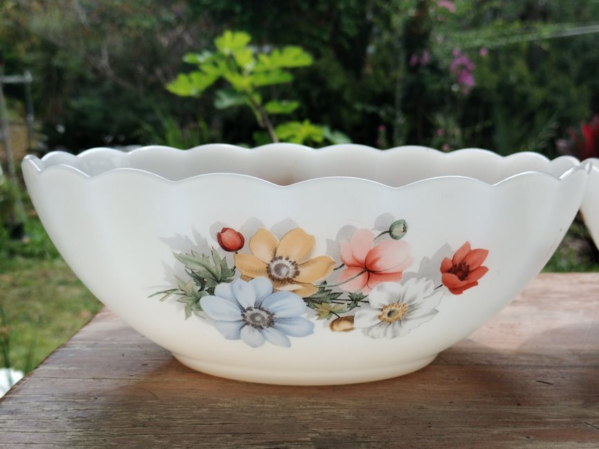 Front view of a scalloped tempered milk glass large salad bowl with multicolour anemone pattern by Arcopal France on a wooden table and a green nature background